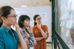 Three women brainstorming and analyzing sticky notes on a glass wall in a meeting room.