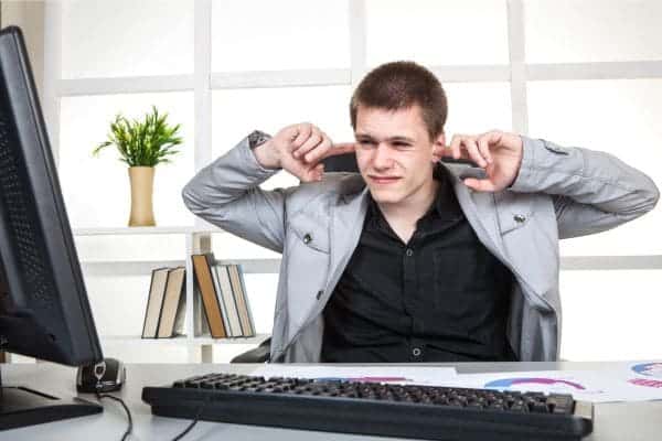 Man at a desk covering his ears in frustration while looking at a computer screen.