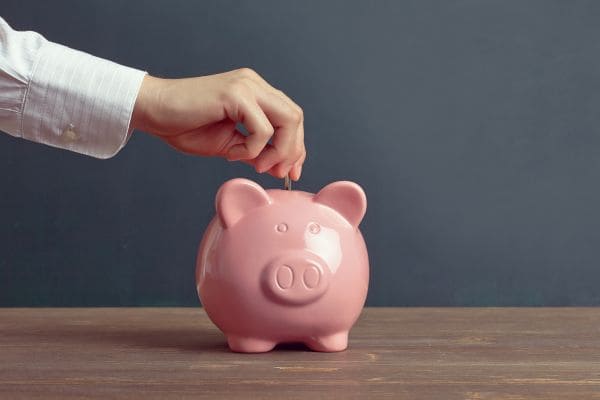 Hand placing a coin into a pink piggy bank on a wooden table.