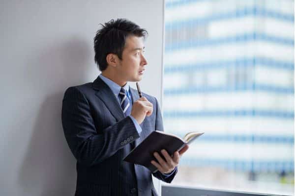 Asian businessman holding a book, gazing thoughtfully out of a window in a modern office setting.
