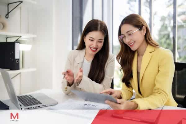 Two professional businesswomen reviewing paperwork and discussing strategy in a modern office workspace.