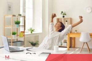 Woman stretching and relaxing at a bright home office desk after working on a laptop computer.