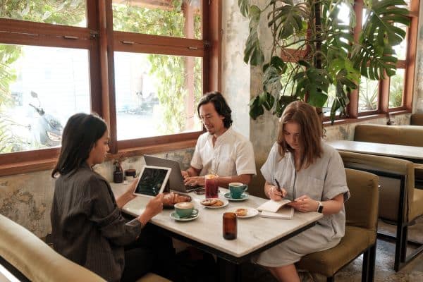 Three coworkers collaborating with laptops and notebooks at a cafe table with coffee and plants
