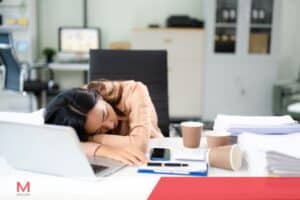 Exhausted female office worker sleeping on desk surrounded by coffee cups and paperwork