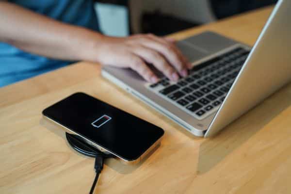 Person using laptop while smartphone charges on wireless charging pad on wooden desk