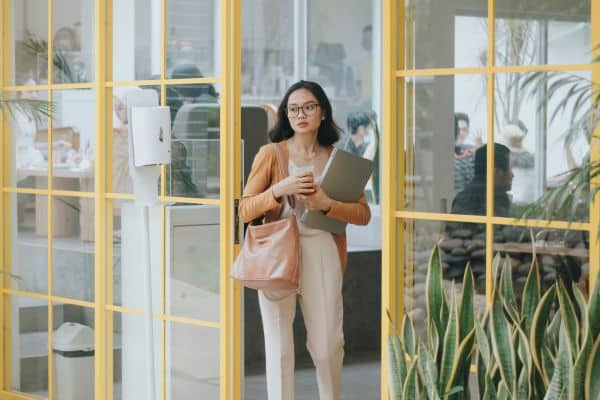 Professional woman in glasses carrying laptop and bag walking through modern office glass doors