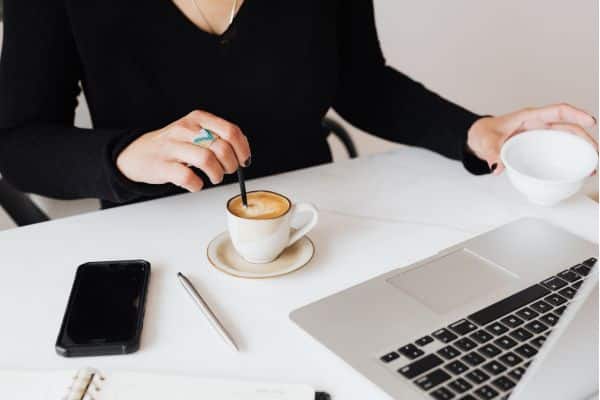 Woman stirring coffee at white desk beside laptop and smartphone in modern workspace