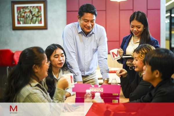 A group of five people seated at a table, sharing coffee from an open box in a casual meeting setting.
